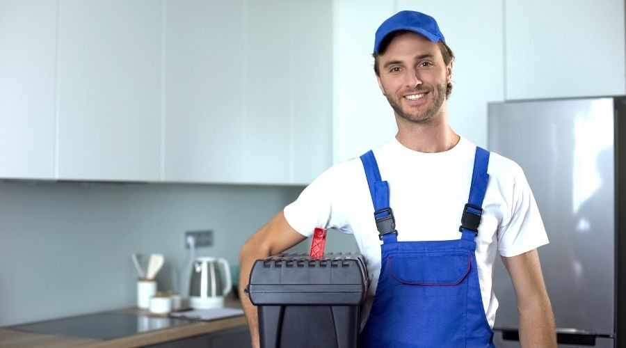 Young plumber in blue overalls and cap holding toolbox in modern kitchen, representing A & W Plumbing and Heating services.