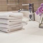 Stacked white bathroom towels beside a vase with a purple flower in a clean sink area by A & W Plumbing and Heating.