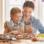A woman and young girl decorate pumpkin-shaped cookies with baking tools on a wooden surface for a Halloween activity.