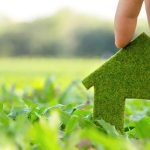 Hand holding a green house-shaped symbol, showcasing eco-friendly plumbing and heating practices with grass background.