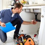 A man kneeling under a kitchen sink, inspecting plumbing with tools, representing plumbing maintenance.