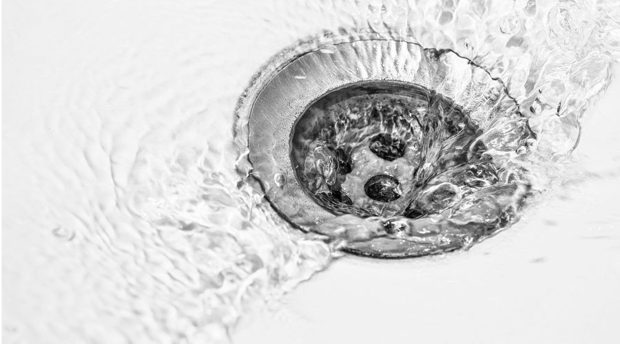 Water flows down a stainless steel drain in a white sink. The water is captured mid-motion, creating a dynamic and fluid effect as it swirls around the drain.