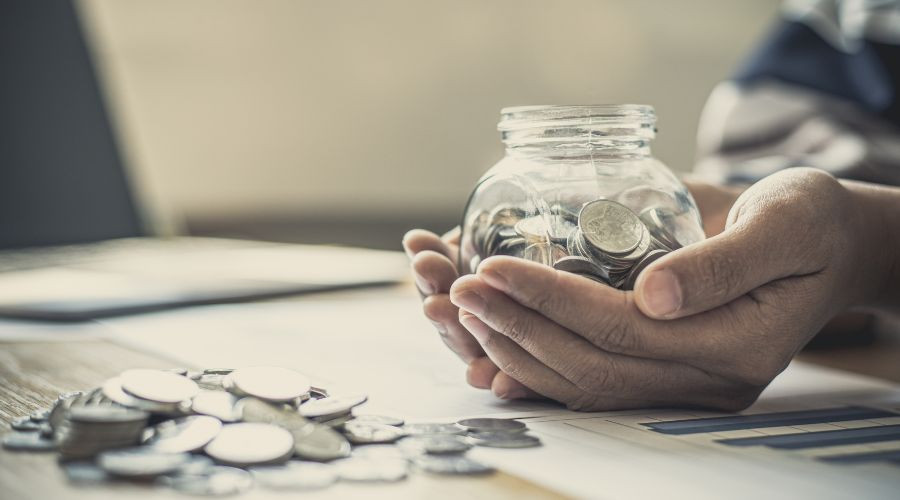 Hands cupping a clear glass jar filled with coins, with more coins scattered on a surface in front of the jar.