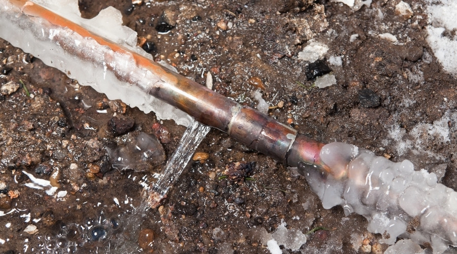 Frozen copper plumbing pipe with ice and water leakage on the ground, illustrating potential plumbing issues during cold weather.
